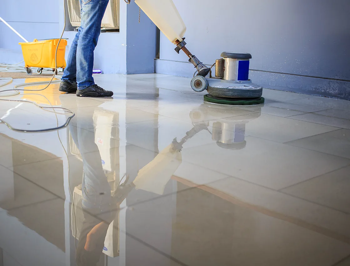 A floor technician running a low-speed rotary floor machine across a glossy commercial tile floor during a strip and wax.
